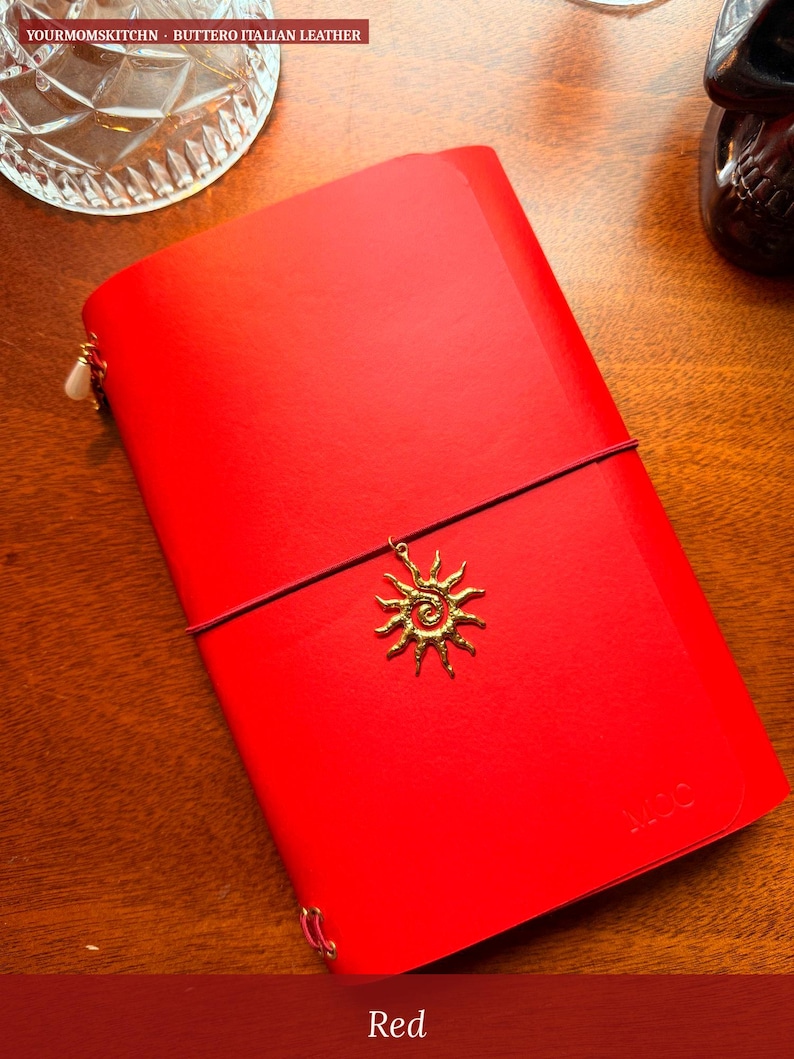 May include: A bright red leather journal with a sun charm and elastic closure. The journal has a gold-colored charm and the word "Red" is at the bottom of the image.