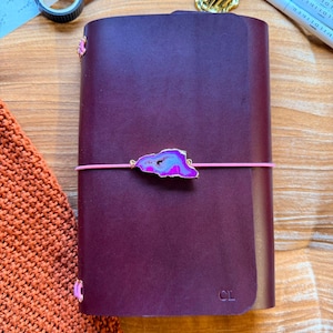 May include: A Bordeaux-colored leather journal with a pink elastic closure and a decorative agate stone accent. The journal has the initials "CL" embossed on the cover. The text "YOURMOMSKITCHN - BUTTERO ITALIAN LEATHER" is visible.
