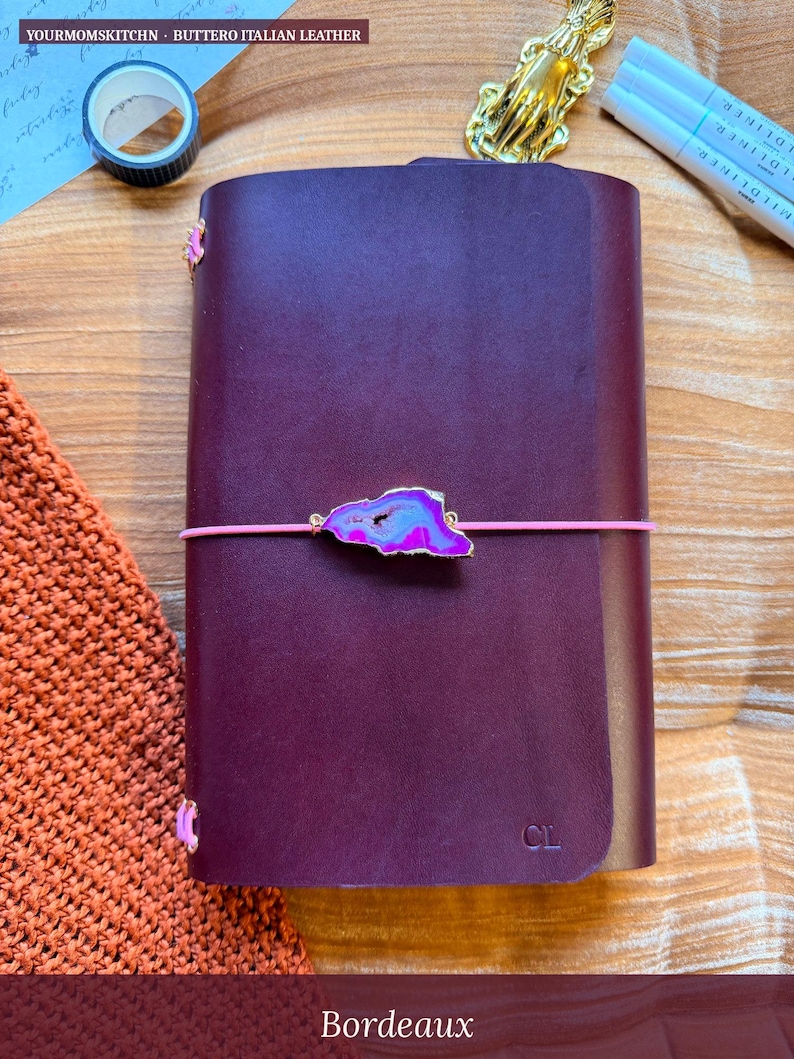 May include: A Bordeaux-colored leather journal with a pink elastic closure and a decorative agate stone accent. The journal has the initials "CL" embossed on the cover. The text "YOURMOMSKITCHN - BUTTERO ITALIAN LEATHER" is visible.