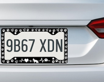 May include: A black license plate frame featuring a white pattern of paw prints and hearts along the border. The bottom edge displays a heartbeat line graphic with a heart, a silhouette of a dog, and a paw print. The plate reads 9B67 XDN.