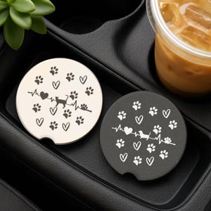 May include: Two car coasters, one cream and one black, with paw prints, hearts, and a heartbeat line design. The cream coaster features a cat silhouette, while the black coaster has a dachshund silhouette. Both are round and designed to fit in a car cup holder.