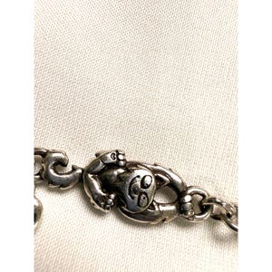 Pode incluir: Close-up de uma pulseira prateada com um berloque de sapo. O sapo tem um rosto sorridente e est&aacute; preso a uma pulseira em estilo de corrente. O fundo &eacute; um tecido de cor clara.