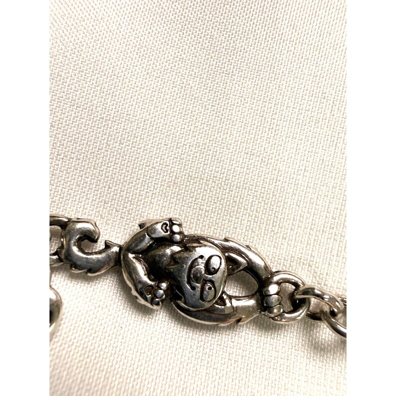 Pode incluir: Close-up de uma pulseira prateada com um berloque de sapo. O sapo tem um rosto sorridente e est&aacute; preso a uma pulseira em estilo de corrente. O fundo &eacute; um tecido de cor clara.