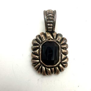 May include: A silver-toned pendant with a rectangular, black gemstone center. The pendant has a decorative, scalloped border and a textured bail for a chain. The gemstone is faceted, reflecting light.