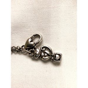 Pode incluir: Close-up de um fecho de colar prateado com elementos de cora&ccedil;&atilde;o e decorativos. O fecho apresenta um fecho em forma de cora&ccedil;&atilde;o e um berloque com um design em forma de cora&ccedil;&atilde;o. A corrente do colar &eacute; vis&iacute;vel.