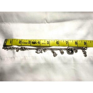 Pode incluir: Uma pulseira de prata com v&aacute;rios berloques, incluindo cora&ccedil;&otilde;es, um rel&oacute;gio, uma fechadura e um cadeado. A pulseira tem aproximadamente 20 cm de comprimento, medida com uma fita m&eacute;trica amarela. Os berloques s&atilde;o detalhados.