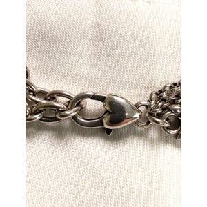 Pode incluir: Close-up de um fecho de colar prateado com um design em forma de cora&ccedil;&atilde;o. O fecho est&aacute; ligado a v&aacute;rios fios de elos de corrente. O fundo &eacute; um tecido claro e texturizado.