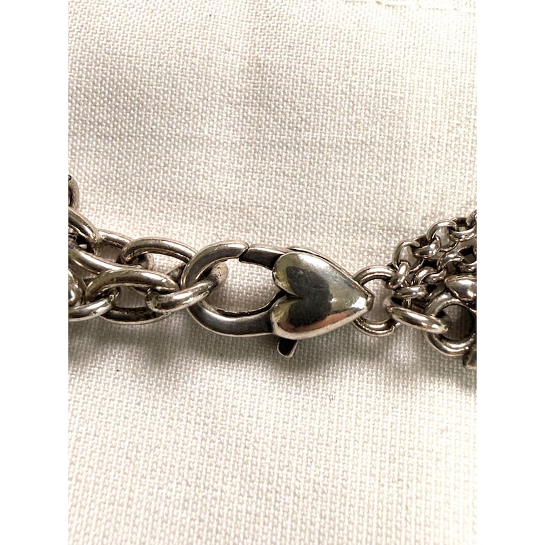 Pode incluir: Close-up de um fecho de colar prateado com um design em forma de cora&ccedil;&atilde;o. O fecho est&aacute; ligado a v&aacute;rios fios de elos de corrente. O fundo &eacute; um tecido claro e texturizado.