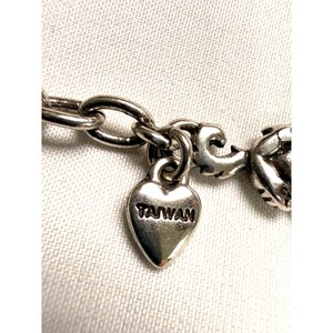 Pode incluir: Close-up de um colar prateado com um pingente em forma de cora&ccedil;&atilde;o. O pingente est&aacute; gravado com a palavra "TAIWAN". O colar est&aacute; sobre um fundo de tecido branco.