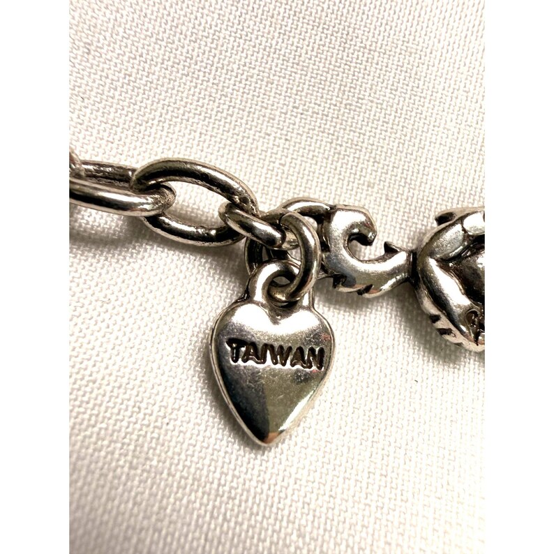 Pode incluir: Close-up de um colar prateado com um pingente em forma de cora&ccedil;&atilde;o. O pingente est&aacute; gravado com a palavra "TAIWAN". O colar est&aacute; sobre um fundo de tecido branco.