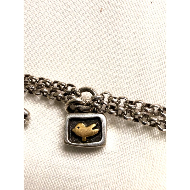 Pode incluir: Colar de corrente prateada com um pingente quadrado com um desenho de p&aacute;ssaro dourado. O p&aacute;ssaro est&aacute; dentro de um quadrado preto, sobre um fundo prateado. A corrente tem um design de dupla corrente.