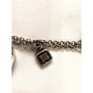 Pode incluir: Close-up de uma pulseira de corrente prateada com um pequeno berloque de cadeado quadrado. O cadeado tem um design dourado com os n&uacute;meros "918". A pulseira est&aacute; em um fundo branco.