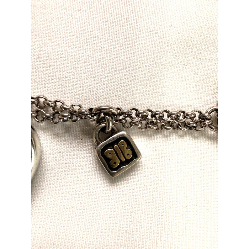 Pode incluir: Close-up de uma pulseira de corrente prateada com um pequeno berloque de cadeado quadrado. O cadeado tem um design dourado com os n&uacute;meros "918". A pulseira est&aacute; em um fundo branco.