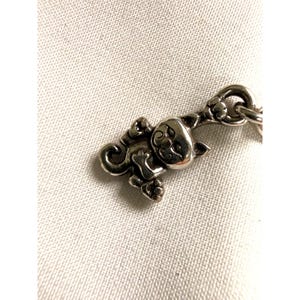 Pode incluir: Um pingente prateado em forma de gato estilizado. O pingente apresenta um design detalhado do rosto, patas e corpo do gato. O pingente &eacute; preso a um elo de corrente. O fundo &eacute; um tecido branco texturizado.