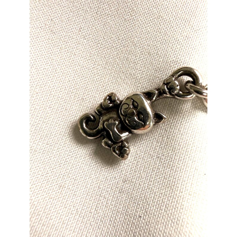 Pode incluir: Um pingente prateado em forma de gato estilizado. O pingente apresenta um design detalhado do rosto, patas e corpo do gato. O pingente &eacute; preso a um elo de corrente. O fundo &eacute; um tecido branco texturizado.