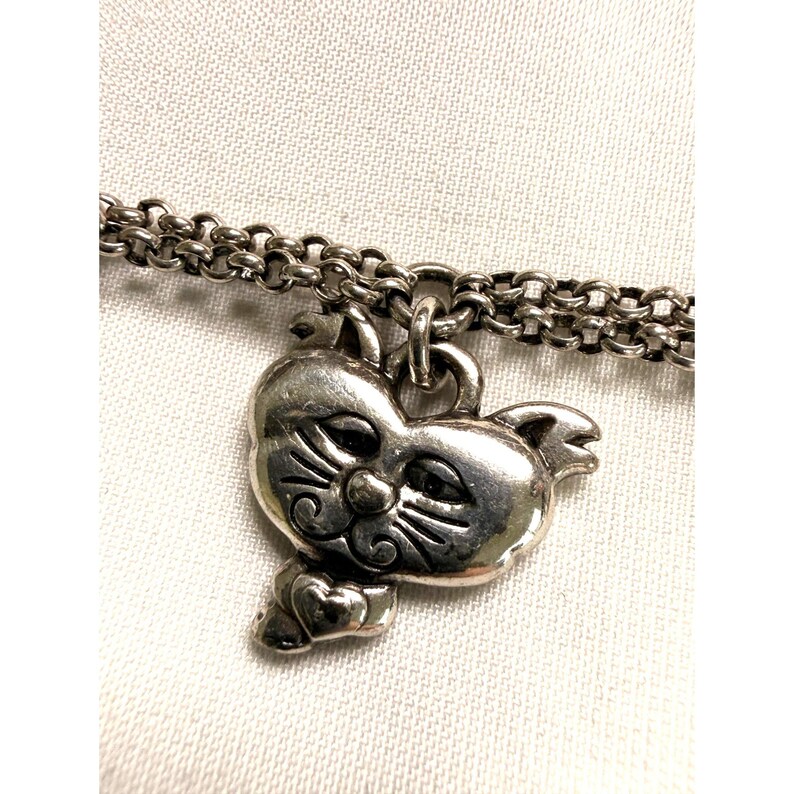 Pode incluir: Uma pulseira com um berloque de gato prateado. O berloque &eacute; um rosto de gato detalhado com bigodes e um la&ccedil;o em forma de cora&ccedil;&atilde;o. A corrente da pulseira &eacute; um design de elos e o berloque pende de um pequeno la&ccedil;o.