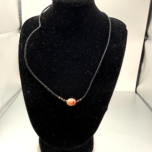 Può includere: Una collana di perline nere con una perla ovale centrale. La perla ovale è principalmente color pesca con strisce rosse e bianche. La collana è esposta su un busto di velluto nero.