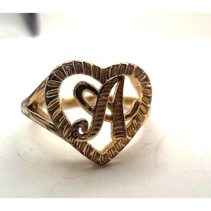 Op de afbeelding: Goudkleurige ring met de letter "A" in een hartvormige lijst. De ring heeft een textuurontwerp rond het hart en de letter. De ring staat tegen een witte achtergrond.