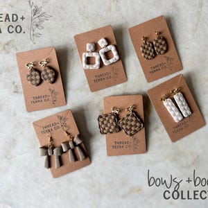May include: A selection of handcrafted earrings presented on brown cards. The earrings showcase woven designs in neutral shades, such as white, brown, and beige. The cards are branded with "THREAD + TERRA CO." and the words "bows + board COLLECTION" are at the bottom.