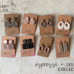 May include: Eight pairs of polymer clay earrings displayed on brown cardboard cards labelled Thread + Terra Co. The collection features various shapes including circles, squares, and arches in neutral tones of beige, brown, grey, and black with textured patterns.