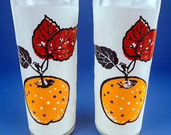 Könnte beinhalten: Zwei hohe, transparente Trinkgläser mit weißem, mattiertem Hintergrund. Jedes Glas zeigt einen leuchtend orangefarbenen Apfel mit weißen Punkten, roten Blättern und schwarzen Blättern. Ideal zum Servieren von Getränken.