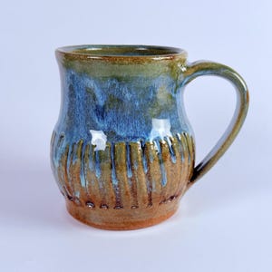 Könnte beinhalten: Eine handgefertigte Keramiktasse mit einer einzigartigen Glasur. Die Tasse hat eine blaue und grüne Glasur auf einem braunen Sockel mit vertikalen strukturierten Linien. Der Griff ist gebogen und die Tasse hat eine breite Öffnung.