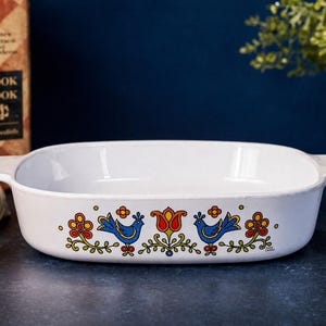 May include: A white rectangular ceramic baking dish with integrated side handles, featuring a colorful folk-art style decal on the front. The design includes two blue birds, a central red tulip, and yellow and red flowers with green stems.