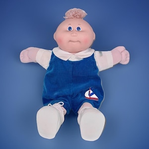 Peut inclure: Poupée Cabbage Patch Kid vintage avec une tête chauve, des yeux bleus et des joues roses. La poupée porte une chemise blanche à col sous une combinaison en velours côtelé bleu avec un écusson de voilier. Des chaussures blanches complètent la tenue.