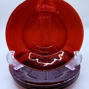 Puede incluir: Una pila de platos de vidrio rojo, cada uno con un soporte decorativo transparente. Los platos tienen un acabado brillante y se exhiben sobre un fondo blanco. Los platos son de un color rojo intenso.