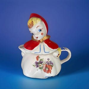 Może przedstawiać: Wazonik na śmietankę z ceramiki w stylu vintage z figurką dziewczynki w czerwonej pelerynie z kapturem. Biały wazonik na śmietankę ma kwiatowy wzór z czerwonymi, fioletowymi i białymi kwiatami. Uchwyt i peleryna dziewczynki są ozdobione złotem.