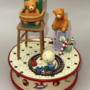 May include: A decorative musical carousel featuring teddy bears, a high chair, and toy blocks. The carousel has a train circling a central ball. The base is decorated with a red trim and toy illustrations.