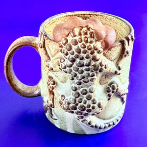 以下が含まれることがあります： 茶色のテクスチャード加工のトカゲのデザインが施されたベージュのセラミックマグ。トカゲはマグの本体を包み込んでいます。ハンドルは茶色で、マグの内側には茶色の雲のような形が入っています。ユニークで手作りの外観。
