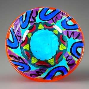 May include: A colorful, hand-painted glass bowl with an orange rim. The bowl features a turquoise interior, a central ring of red, green, and yellow, and blue and purple abstract shapes. The design is bold and vibrant.