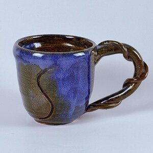 May include: Handmade ceramic mug with a unique twisted handle. The mug features a deep blue glaze on the exterior, transitioning to a brown and green base. The interior is a rich brown color, perfect for coffee or tea.