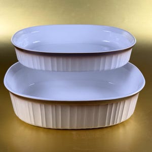 May include: Two oval, off-white ceramic baking dishes stacked. They have a light blue interior and a brown rim. The exterior features vertical ribbed detailing. Ideal for baking and serving.