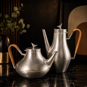 May include: Two silver teapots with woven handles and bird-shaped lid ornaments. One teapot is round, the other is tall and slender. The teapots are displayed on a black marble surface, with a dark background and decorative elements.