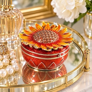 May include: A round, red enameled trinket box featuring a three-dimensional sunflower lid with orange and yellow petals. The base of the box is decorated with a repeating triangular pattern in gold, and the lid is hinged to the main body.