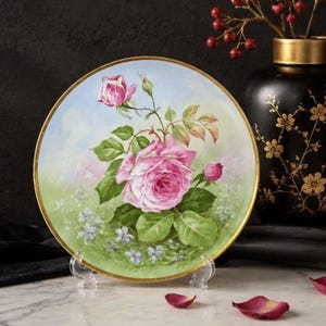 May include: A hand-painted decorative plate featuring a pink rose with green leaves and buds. The plate has a gold rim and is displayed on a clear stand. The background has a light blue sky and green grass with small purple flowers.