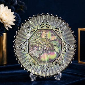 Puede incluir: Un plato de cristal iridiscente de forma redonda con un panel hexagonal central en relieve que muestra un detalle de uvas, hojas y frutas. El plato tiene un borde festoneado y dentado con un patrón geométrico que irradia desde el centro.