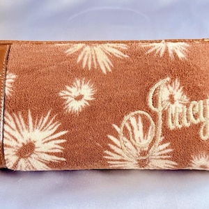 May include: A brown wallet featuring a floral design and the word "Juicy" embroidered in cream thread. The wallet has a brown patent leather accent and a gold zip.