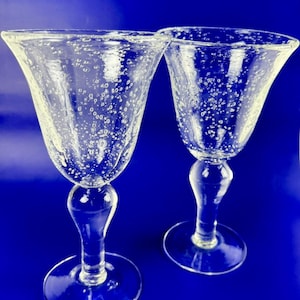 May include: Two clear, stemmed wine glasses with a flared bowl design. The glass has a bubbly texture throughout. The glasses sit on a round base and have a curved stem. The background is a solid blue.