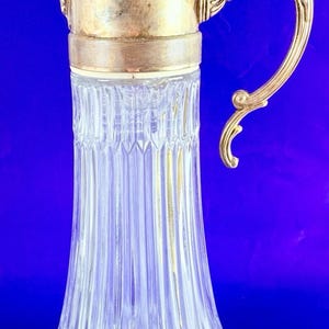 May include: A vintage clear glass pitcher with a gold-coloured metal lid, handle, and accents. The pitcher has a ribbed design and a decorative handle. The lid has a small finial on top. The background is a solid blue.