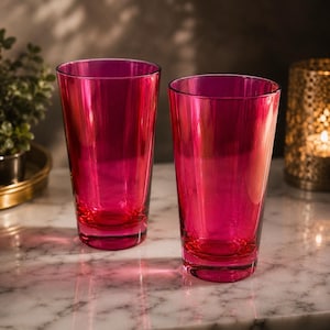 May include: Two tall, vibrant magenta-colored drinking glasses. The glasses are made of transparent glass and have a simple, elegant design. They are placed on a marble surface, with a plant and a candle holder in the background.