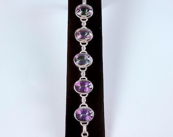 Peut inclure: Un bracelet en argent orné de cinq pierres précieuses ovales et irisées. Les pierres passent du vert au violet profond. Le bracelet est relié par des maillons en argent et possède un fermoir à bascule. Le bijou est présenté sur un support en velours noir.