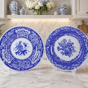 May include: Two blue and white decorative plates with floral patterns. The plates are presented on clear stands, highlighting the detailed designs and a traditional style. Each plate has a central floral design and decorative borders.