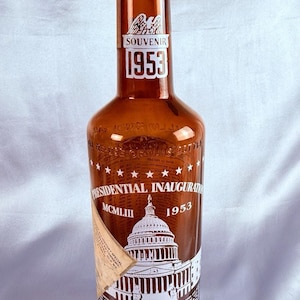 May include: A brown glass souvenir bottle from the 1953 Presidential Inauguration. The bottle features a label with the Capitol building, stars, and the text "SOUVENIR 1953" and "PRESIDENTIAL INAUGURATION MCMLIII 1953".