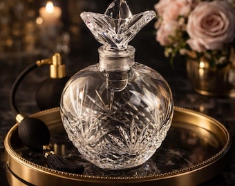 May include: A clear, cut-glass perfume bottle with a starburst design and a decorative stopper. The bottle sits on a gold-rimmed tray, with a black atomizer and a bouquet of pink roses in the background.