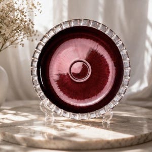 Puede incluir: Un plato circular de vidrio con un centro de color burdeos intenso y una textura radial similar a un rayo de sol. El plato tiene un borde ancho, transparente y facetado con un patrón rectangular repetido, sostenido por un soporte.