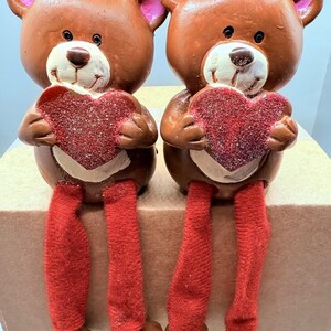 May include: Two brown bear figurines with pink inner ears, white muzzles, and black noses. Each bear holds a glittery red heart and features long, dangling red fabric legs. The bears have a smooth finish and are positioned side by side.