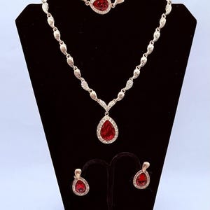 May include: A jewelry set consisting of a necklace and matching earrings. The necklace features a teardrop-shaped red stone pendant surrounded by clear crystals, suspended from a chain of textured silver-tone links. Two matching teardrop earrings.